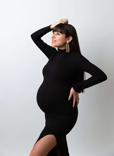 Rochel Konik Photography | Top  Brooklyn NY Maternity Photographer captures expectant mom in fitted black dress with one hand under her bump, the other resting on her back. Mom is looking up at the light, smiling. Images is shot side-profile.