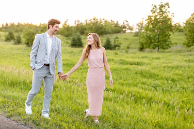 A white couple walking in the grass. He is wearing a gray suit and she is wearing a blush dress.