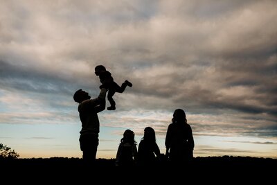 Silhouetted family enjoying a moment outside, with a child being lifted into the air against a cloudy evening sky, capturing a beautiful scene for photography in Pittsburgh PA.