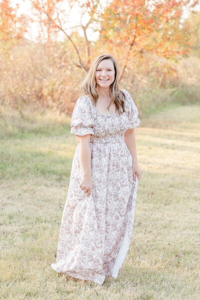 A women in a beautiful neutral color dress with flowers stands in a field posing for the camera.
