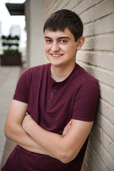 Senior boy in red shirt smiles against wall.