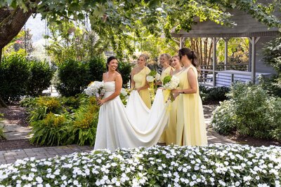 A bride in a strapless white gown walks through a lush garden surrounded by her bridesmaids in soft yellow dresses. They carry white bouquets while helping to hold her dress train, all sharing laughter beneath the shade of green trees and blooming flowers.