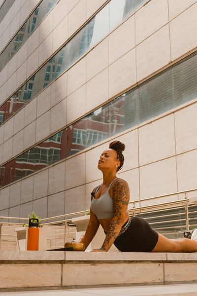 Woman instructor practicing yoga outdoors at a Yoga and Mimosas event in Houston, Texas, performing a stretching pose with a mimosa on the side in a modern urban setting.