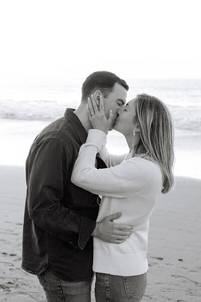 black and white image of engaged couple kissing on the beach.