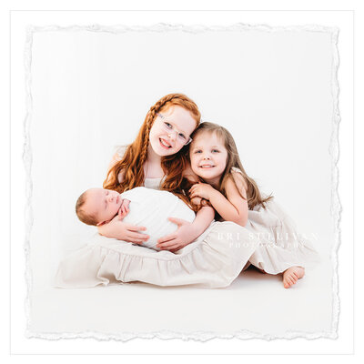 The photo shows a sweet moment between a 5-year-old boy and his new baby sister. The boy sits on an all-white floor, holding his sister in his arms. The background is a clean and pure white studio, creating a classic and elegant setting. Shot by photographer Bri Sullivan, this photo captures the warmth of sibling love.