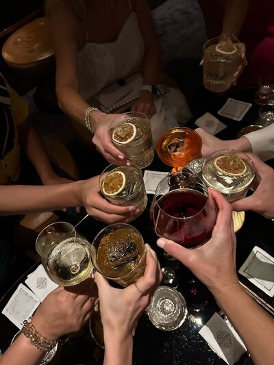 A group of people toasts with various drinks, including wine and cocktails garnished with dried fruit, in celebration. The photo is taken from an overhead perspective, capturing the moment of cheers