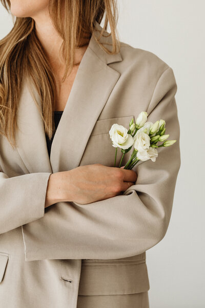 A woman in a beige blazer crosses her arms, holding a small bunch of white flowers in one hand. Her ginger hair frames her shoulders.