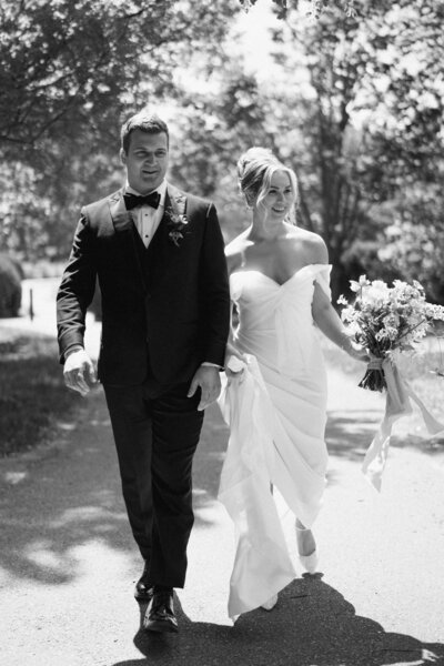 Black and white photo of a bride and groom walking outdoors, with the bride in an off-the-shoulder gown holding a bouquet and the groom in a classic tuxedo.