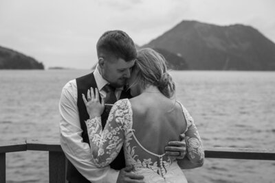 Newlyweds paddle a bright green kayak through the waters of coastal Alaska, celebrating with a bouquet and paddle raised. A scenic footbridge and a group of cheering guests stand on a wooden deck above, with lush mountains in the background, capturing the adventurous spirit of an Alaska elopement.