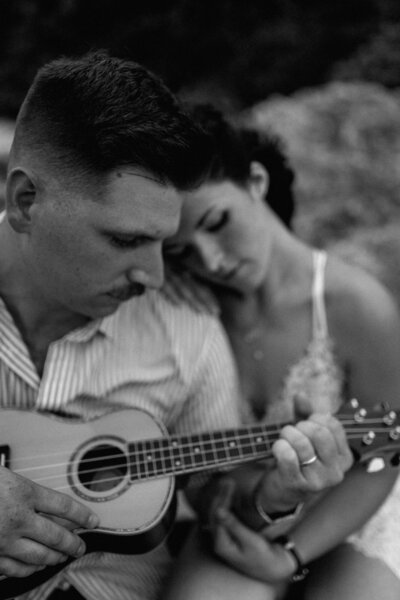 M is playing the ukelele while N rests her head on his shoulder and one hand rests on his leg holding his arm gently