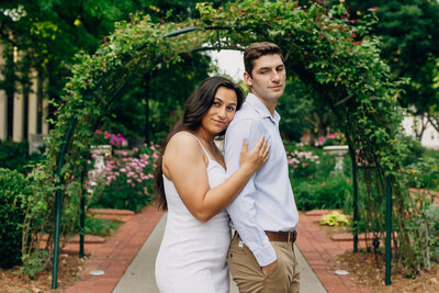 Woman gently leaning on her fiancé’s back while he looks over his shoulder at her with a soft smile.