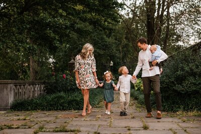 A family of four, captured by a Pittsburgh family photographer, walking hand in hand along a cobblestone path in a park.