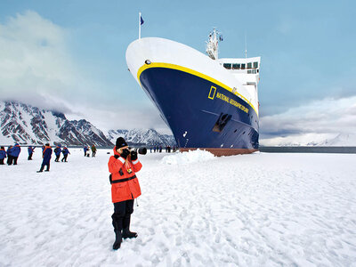 A photographer in an orange parka takes a picture in front of a large blue and yellow expedition ship, “National Geographic Explorer,” on an icy landscape.