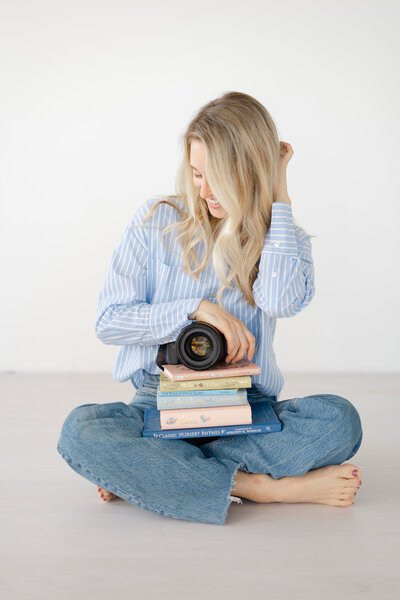 a woman sits cross-legged on the floor with a white background as she tucks her hair behind her ear while holding her photography camera and a stack of children's storybooks