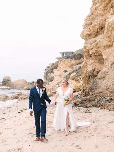 laguna beach elopement of bride in a flowing white dress and groom in a navy suit walk hand in hand on a sandy beach, with rocky cliffs and ocean in the background.