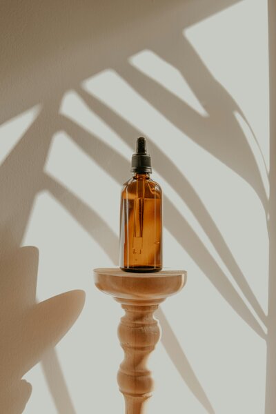 A brown glass dropper bottle rests on a stone-textured pillar against a beige background. Palm leaf shadows create a fresh, natural ambiance.
