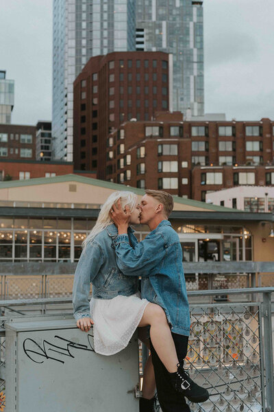 Couple wearing denim jackets kisses on rooftop in front of downtown cityscape