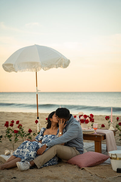 couple share a kiss with a beach picnic set up, styled proposal setup by Yours Truly, a wedding planner and designer in New Jersey.