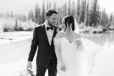 Black and white photo of a bride and groom smiling at each other while walking by the Bow River in Canmore Alberta on a warm summer day after their wedding ceremony