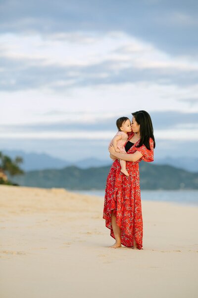 A woman wearing a red dress kisses her baby on the beach.