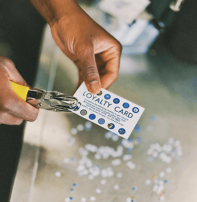 In a bustling Texas coffee shop, a person uses a handheld hole punch to add another mark to their loyalty card. The card, adorned with blue and white symbols, hints at the cozy haven where it began. Confetti from previous punches lies scattered like tiny celebratory reminders on the metal surface below.