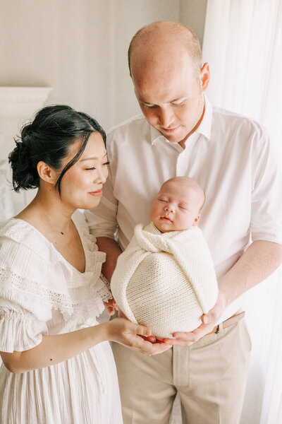 dad holding son in their home being photographed. wife is standing next to him and also wearing white and smiling at son