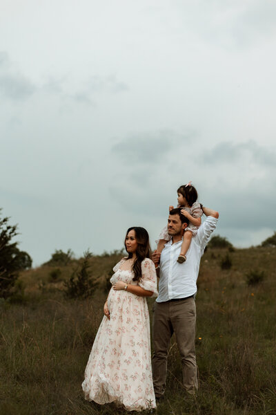 Pregnant woman smiling while her husband holds toddler daughter on his shoulders