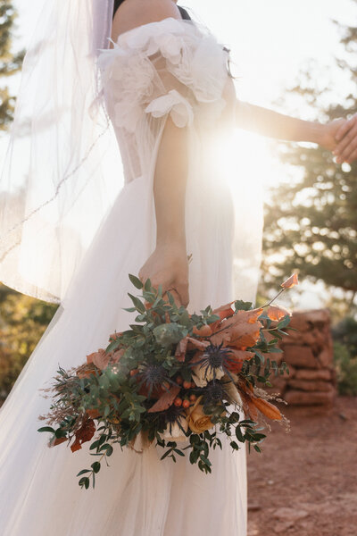 Close-up of bride holding a fall-inspired bouquet with sunset light shining through at her Sedona elopement at Merry Go Round Rock.