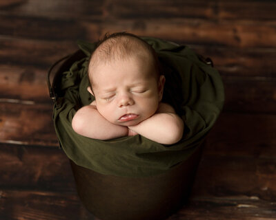 Newborn boy sleeping in a metal pail with his chin resting on his arms. Wood background and army green wrap inside the pail.