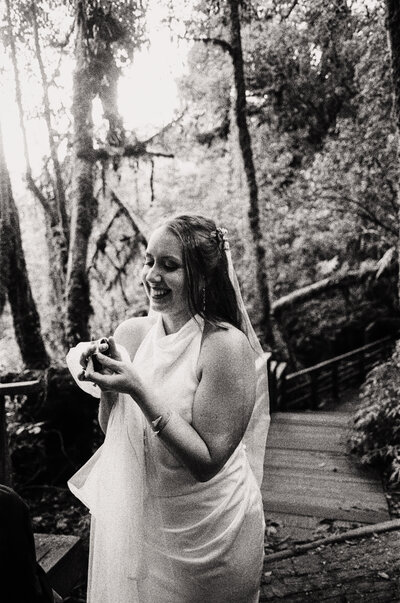 Black and white film photo of a bride smiling while adjusting her ring in a misty rainforest setting during her Thailand elopement — an intimate moment made by a film wedding photographer.