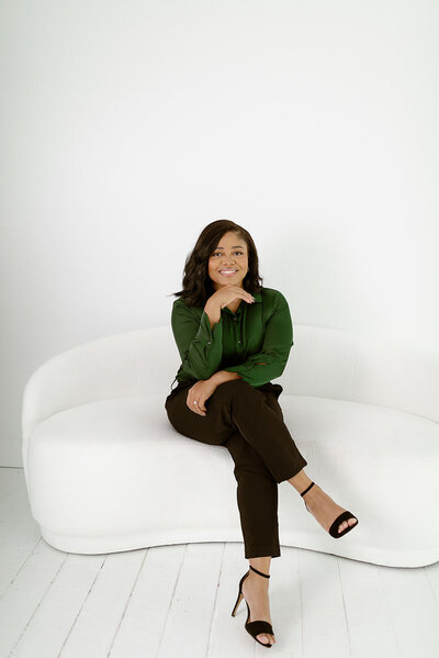 A woman seated confidently on a modern white couch, wearing a green blouse and black pants with black heels. She is smiling with her hand resting under her chin, creating a poised and approachable look in a minimalist setting.