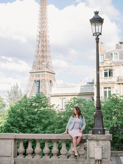 a woman in a jean jacket and pastel dress sitting on a ledge next to a lamp post with the eiffel tower behind her