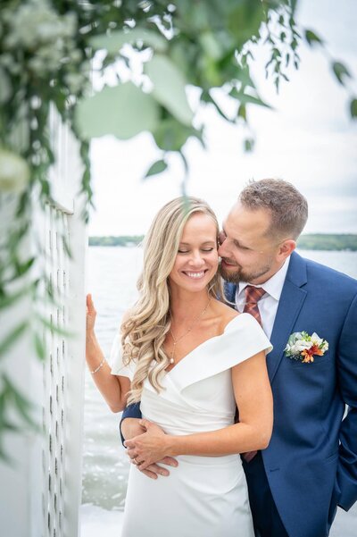 groom whispers in bride's ear neck to trellis in front of lake
