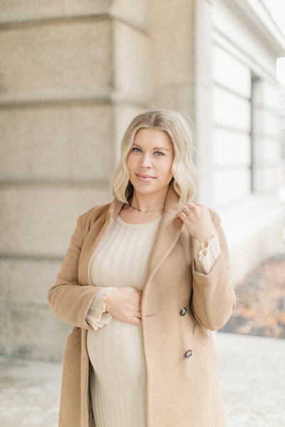 A glowing expectant mother standing outdoors, wearing a beige coat and a cream sweater dress, looking serene against a softly lit stone building backdrop in Kansas City 