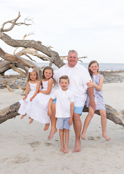 grandfather photographed with grandchildren on a tree on driftwood beach on jekyll island