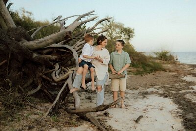 A woman stands on a sandy beach near driftwood, holding a young boy while talking to an older boy who holds a basket. Trees and water are visible in the background.
