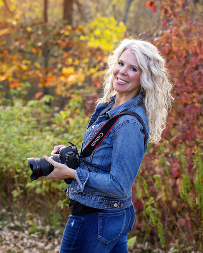 The owner of Studio 64 Photography is holding her camera outside with the fall colors surrounding her smiling back over her shoulder.