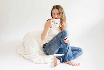Portrait of woman holding a white cup with blue birds on it in one hand while smiling and sitting down on a cream rug.