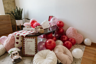Adorable Valentine’s Day photoshoot backdrop using a wood coffee table rented from Beautifully Layered Event Rentals in Milwaukee covered in red, pink and white balloons, vases and pillows
