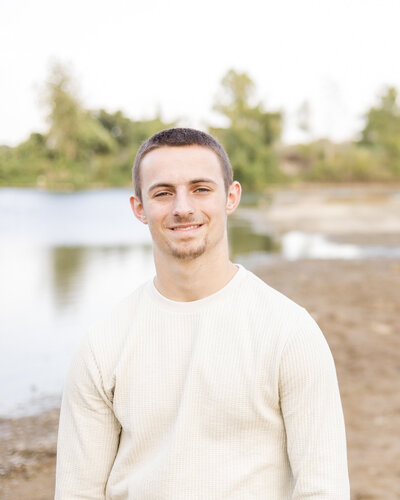 high school senior male standing next to a pond with trees behind him, looking at the camera smiling, with short brown hair and a long shirt