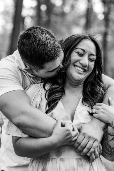 Black and white image of Robert hugging Alexa around her shoulders, she’s holding his hands, smiling at the camera while he kisses her neck.