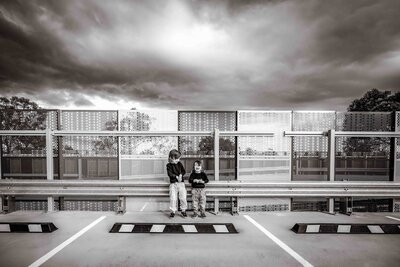 Black and white image of two little boys laughing at each other taken at a roof top carpark with epic skies