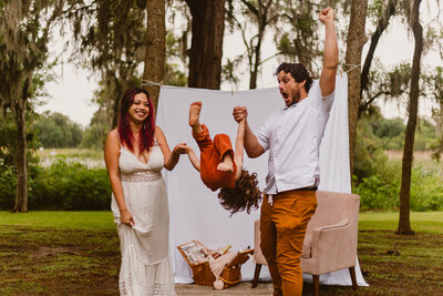 Family celebrating Mother’s Day at Bay Area Park in Texas with a playful picnic setup,  a white sheet background while swinging their young child upside down, all smiling and enjoying the moment. Surrounded by mossy trees