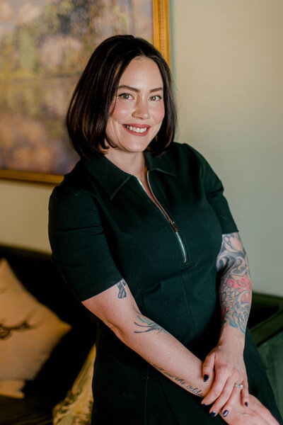 A woman with shoulder-length dark hair smiles, standing indoors. She is wearing a dark short-sleeve dress with a zipper and displays tattoos on her arms. A painting and cushions are visible in the background.