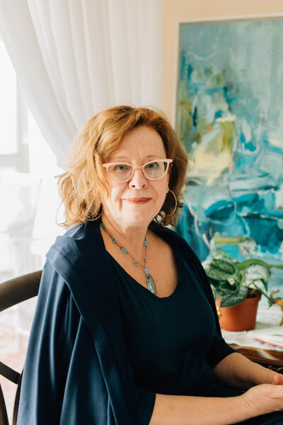 photo of owner Barbara sitting in blue sweater with painting behind her.