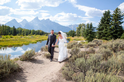 Bride and groom stroll through Schwabacher Landing, surrounded by lush scenery and the Grand Tetons, creating a picturesque Jackson Hole wedding moment.