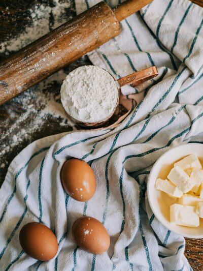 Eggs, butter, a cup of flour, and a rolling pin laying on a towel with flour scattered around