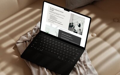 Open laptop resting on a beige couch, displaying a website with a black-and-white photo of kaya ramjee drinking a protein shake and text about elite performers' struggles and success traps.