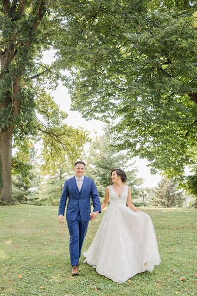 A happy couple holding hands and walking in a park, the woman in a flowing white wedding dress and the man in a blue suit, beautifully coordinated by an Illinois wedding planner.