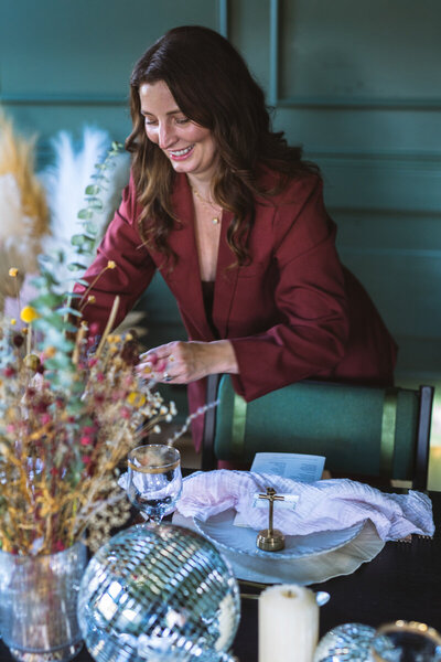 A woman in a red blazer, possibly an event planner, arranges a stylish table setting with dried flowers, a disco ball, and glassware, showcasing her expertise in luxury event planning in Northwest Arkansas.